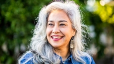 Person with long gray hair smiling while standing in front of trees