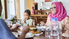 Three people sitting around a table with food on it. One person is wearing a pink head scarf, one a blue shirt and the other a white shirt