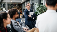 A person standing up, holding paperwork watching something while two other people are sitting and one other person has their back turned