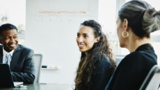 Three people sitting around a table with one smiling and the other two looking at the one smiling