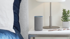 Light gray Amazon Alexa device on a bedside table next to a lamp, watch and houseplant