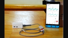 Amrita Spandanam featuring a finger clip and a mobile app showing vital parameter measurements