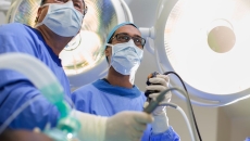 Two surgeons looking down at a camera with scrubs on while holding surgical tools