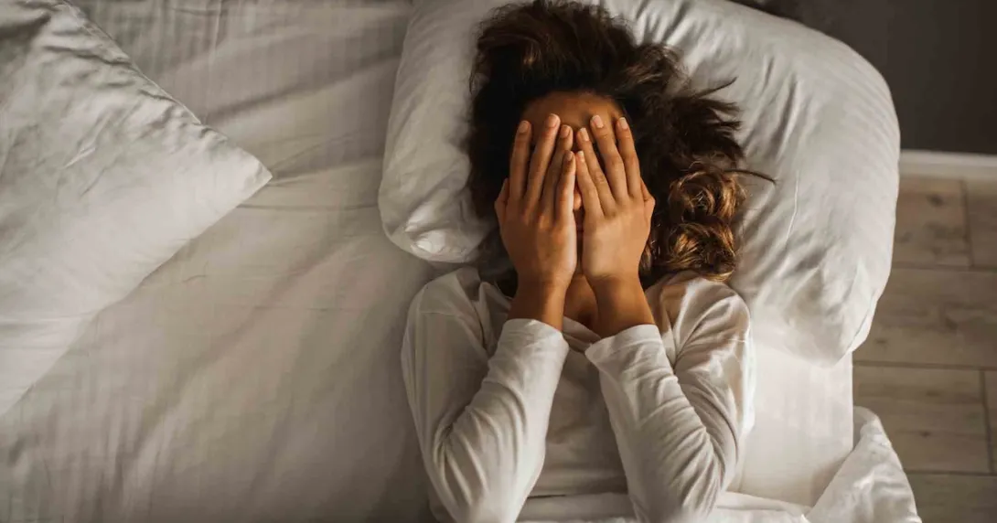 Person lying in bed with their hands over their eyes