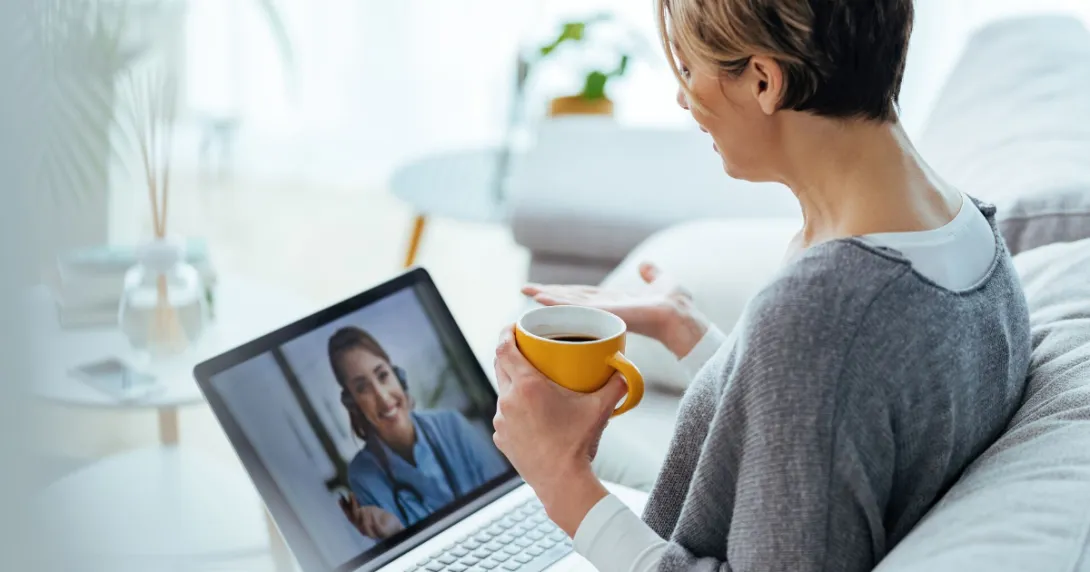 Person sitting on a couch speaking to a healthcare professional on a computer while holding a cup in their hand