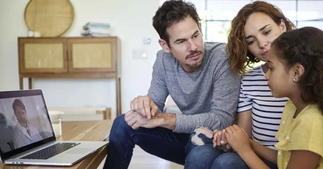 Three people sitting on a couch with a computer on a coffee table showing a healthcare provider on the screen