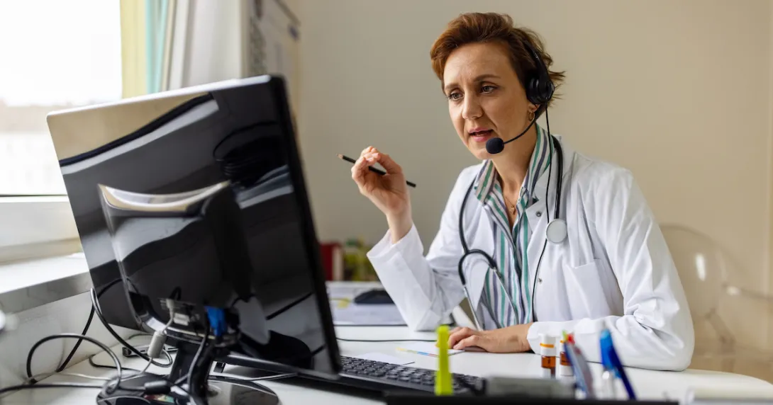 Healthcare provider sitting at a desk wearing a lab coat and headphones while talking to someone on a computer