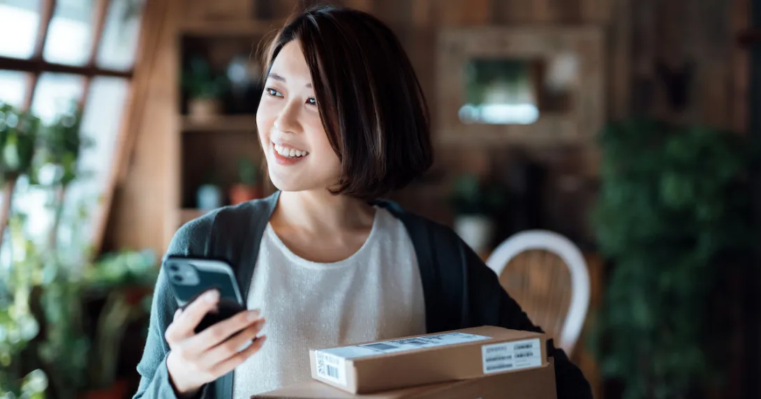 Person standing in their living room holding a phone and standing by stacked boxes