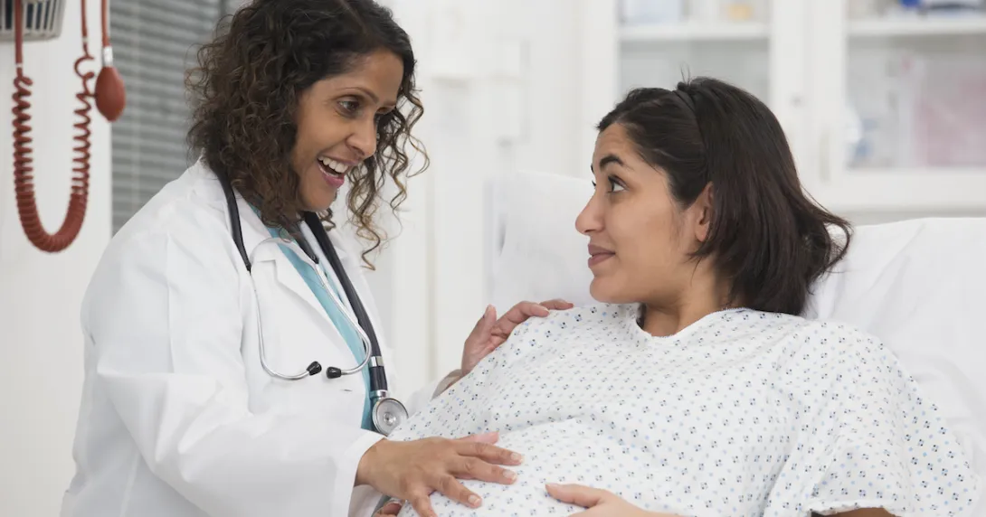 Healthcare worker standing beside a pregnant person lying in bed while both are touching the pregnant person's stomach