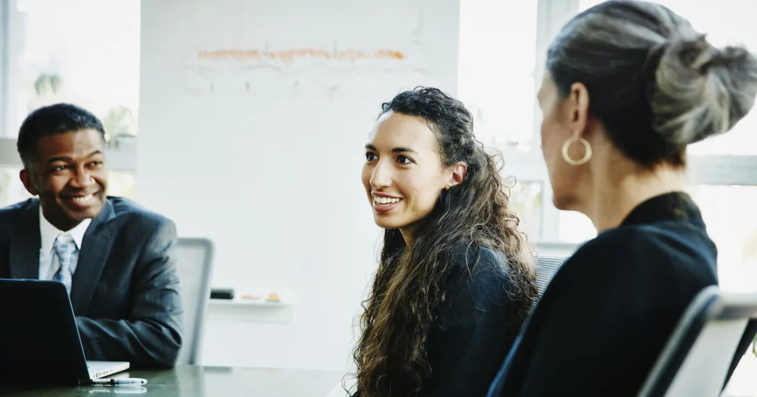 Three people sitting around a table with one smiling and the other two looking at the one smiling