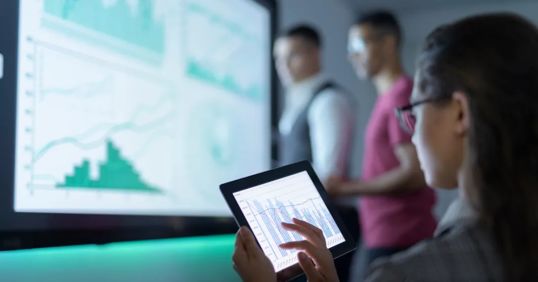 Person sitting while looking at a tablet with two other people standing up looking at a graph on a screen