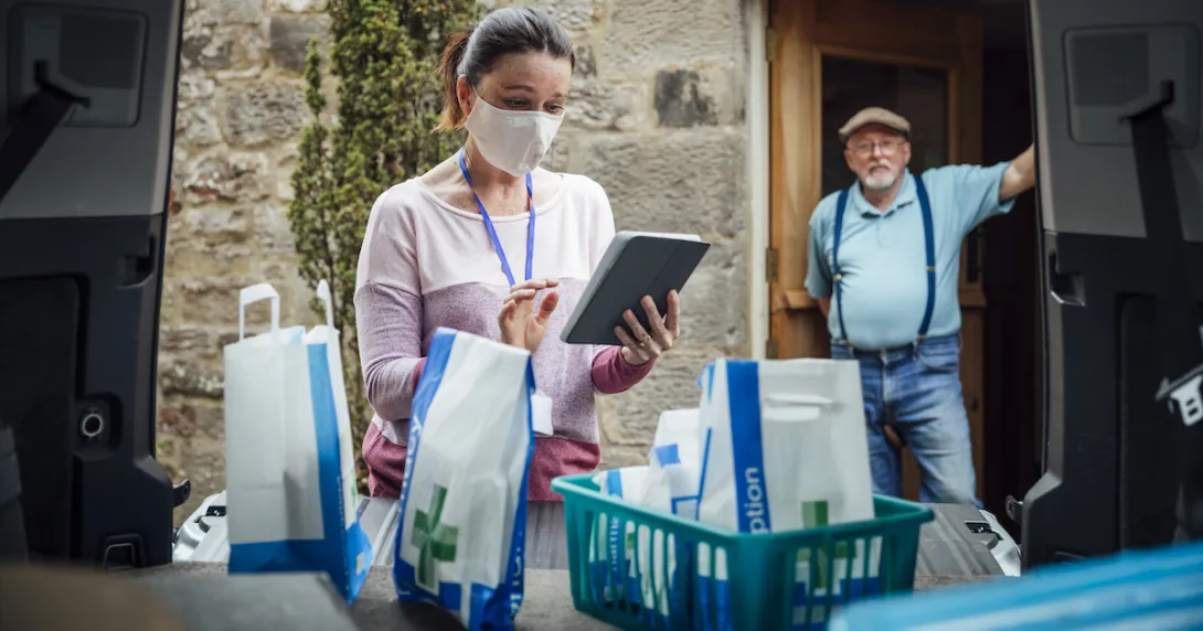 A woman using a tablet delivering medications while a man stands in his doorway in the background