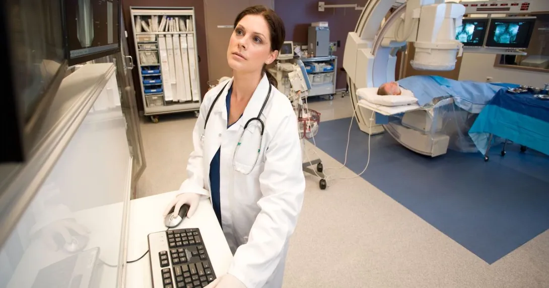 A medical professional looks at images while a patient receives a CT scan in the background