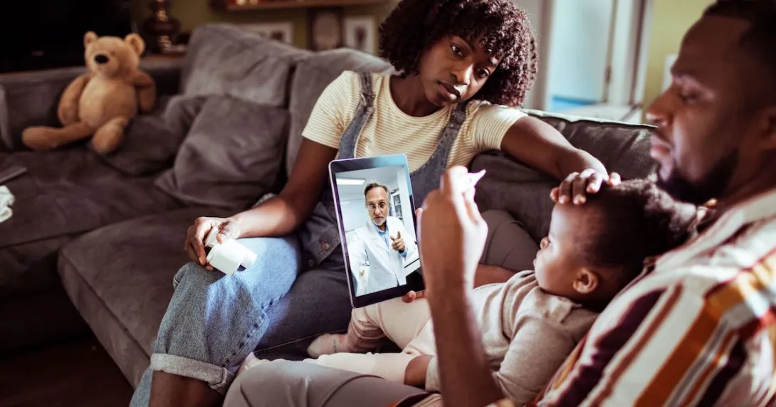 Two people sitting on a couch with a child sitting on one's lap while talking to a healthcare provider on a computer