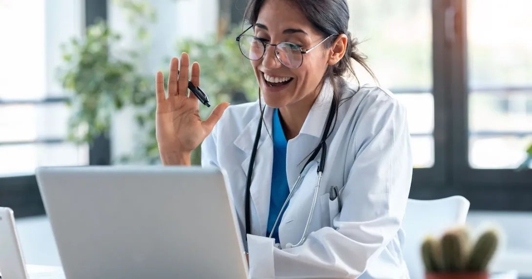 A healthcare provider in a lab coat with a stethoscope around their neck sitting at a desk looking at a computer and waving