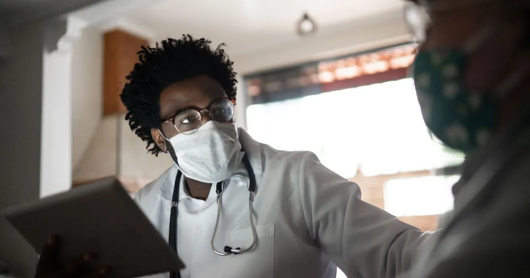 Healthcare provider wearing a mask, a lab coat and a stethoscope around their neck talking to a patient in their home