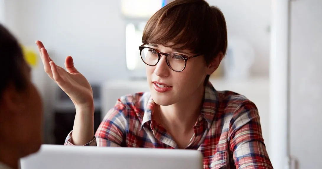 A person with a red plaid shirt on with short hair wearing glass, sitting at a computer with their hand up while talking
