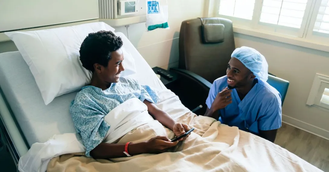 Healthcare provider sitting next to a hospital bed with a teenager in it looking at a phone